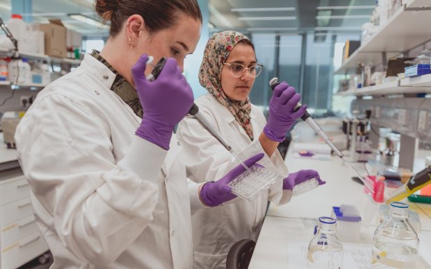 Two researchers in white coats and purple latex gloves are working at a lab station