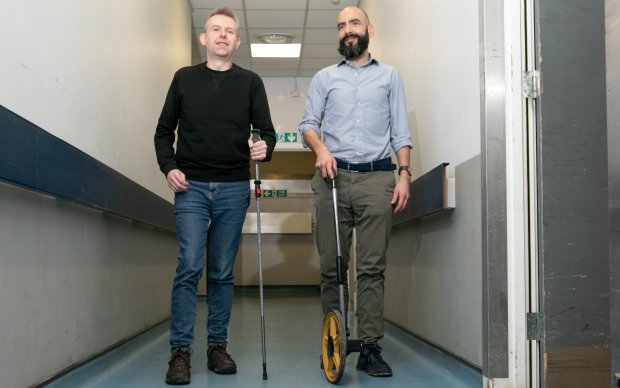 Two men are in a corridor. The man on the left wears a black jumper and jeans, and uses a walking stick. The man on the right has a beard and wears a shirt, he is using a measuring wheel.