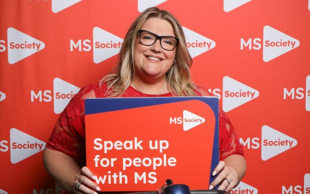Kerry has long blonde hair and wears glasses, she holds up a sign reading "Speak up for people with MS"