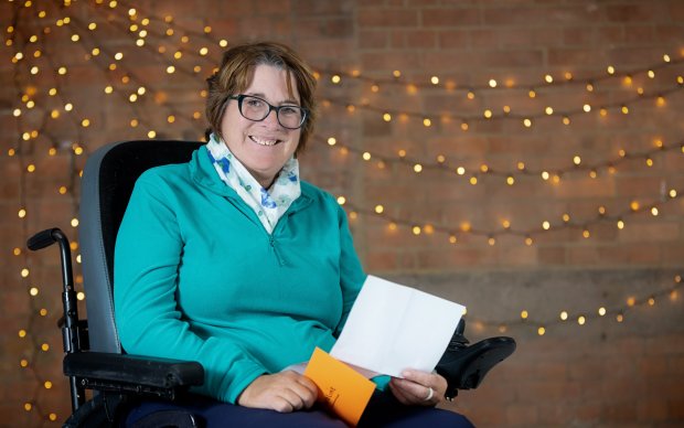 A woman in a turquoise jumper and glasses, holding a piece of paper, sat in a wheelchair
