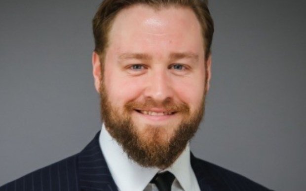 A headshot of a man in a suit with a dark beard against a grey background