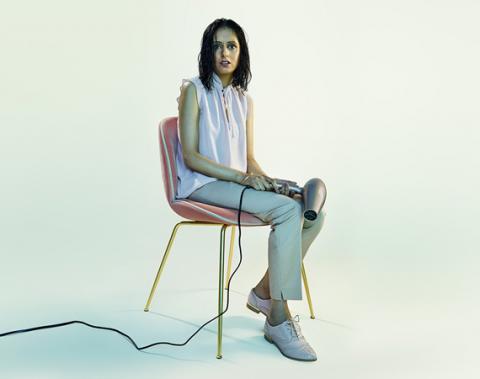 Woman with wet hair sits on a chair holding a hair dryer and looking to camera