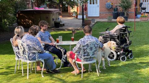 Delia, family and addtional friend sit around a table on the lawn, looking back towards the house. They are joined by a very fluffy dog.