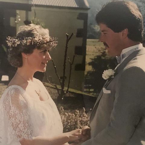 Robyn on her wedding day with short brown hair in white wedding dress holding hands with Grant who has dark hair and a moustache wearing a grey suit.