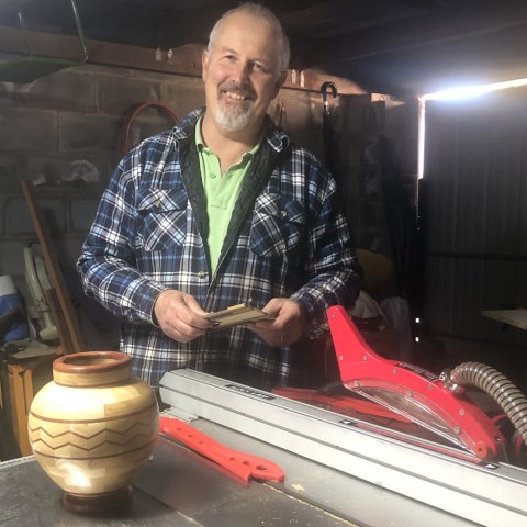 Grant wearing a plaid shirt smiling with a wooden vase he's made on his lathe