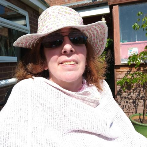 Portia who has advanced MS wearing dark glasses and a white straw hat and shawl in her garden