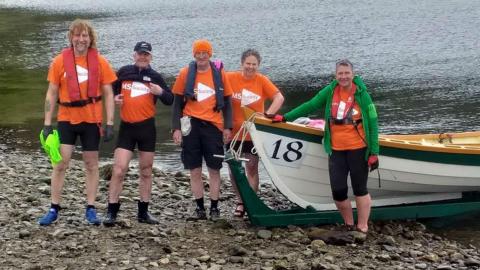 The crew of Rowing the Minch for MS standing by their boat on a pebbly beach