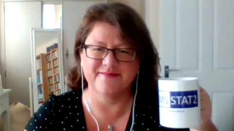 Libby sits at home wearing headphones and holding up a mug saying 'STAT-2'