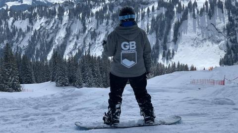 Nina stands on her snowboard facing away from the camera to a view of a snowy, rocky mountain and fir trees.