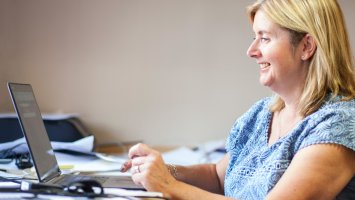 Photo: a woman sat at a desk looking at the MS forum on a laptop she is smiling