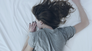 Woman lies face down sleeping on white sheets, she is wearing a grey t-shirt with hair splayed acreoss the bed