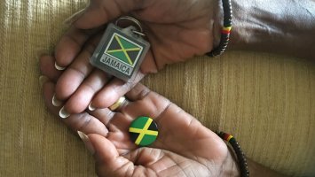 Image shows the hands of Karen's mum, holding a Jamaican badge and key ring and wearing bracelets showing Jamaican colours. 