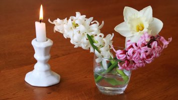 White candle burns in a china holder next to a glass of spring flowers