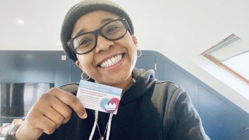Janade smiling to camera wearing a black beanie hat and glasses holding her COVID-19 vaccine card