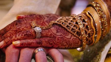 Manicured hand wearing mehndi designs and gold jewellery rests on a pale hand with a ring.