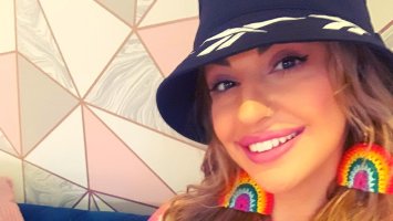 Yazzie smiles wearing crotched rainbow earrings and a black floppy hat.