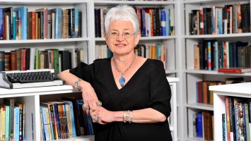Jacqueline Wilson in front of bookshelves, leaning against shelf with typewriter on it