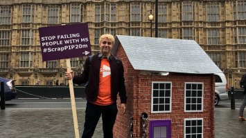 A man in an MS Society t-shirt is standing outside the Houses of Parliament holding a sign that reads "Stop failing people with MS #ScrapPIP20m"  he is stood next to a model house with a chain slung through it. 