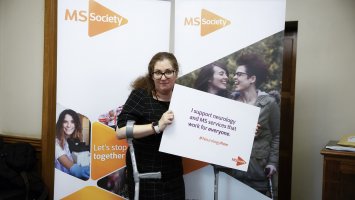 A woman holding a neurology now placard is stood in front of two MS Society banners