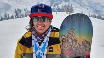 Snowboarder Nina Sparks stands with her board next to her on the snow, wearing a baseball gap and sunglasses, with 3 medals round her neck. 