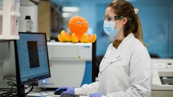 A researcher in a lab coat wearing a mask stands using a computer, with an orange cuddly octopus perched on a shelf beside her