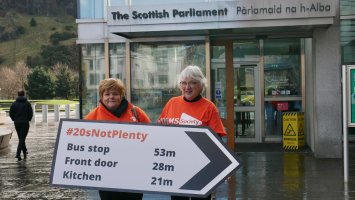 Two women in orange MS Society t-shirts are holding a road sign shaped placard reading #20sNotPlenty. They're stood outside The Scottish Parliament