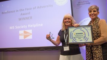 Two women stand holding a framed certificate with the word Winner on it. The woman to th eleft also holds up a small glass trophy in the shape of a star. Behind them is a large screen reading:  Resilience in the Face of Adversity Award Winner, MS Helpline