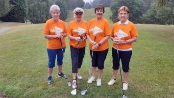 Four women stand side by side in orange MS Society t-shirts on a golfing green, each holds a golf club in front of them