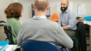 Three people sit in a hospital consulting room. A trial participant is sitting in a wheelchair. Her husband has his back to the camera. A researcher with a beard is smiling at the. An orange fluffy octopus is on a desk behind him. 