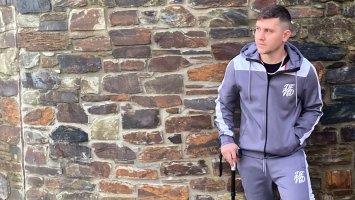 Robbie, wearing a tracksuit and using a walking stick, stands against a stone wall.
