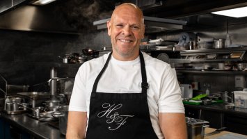 Tom is standing in his restaurant's kitchen wearing a white t-shirt and a black apron. He's smiling at the camera with his arms down by his sides.