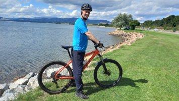 Neil MacLeod stands astride his bike at the edge of a lake, with mountains 