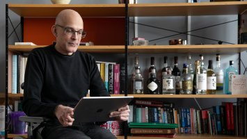 A man in a wheelchair is holding a tablet, behind him is a book case with many books and spirit bottles