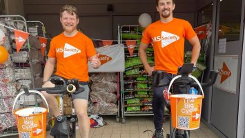Two young men sat on static bicycles, smiling and wearing MS Society T shirts, with MS Society fundraising buckets over the handlebars.