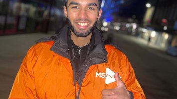 A fundraiser in an orange MS Society jacket stands on a dark street, smiling with thumb raised.