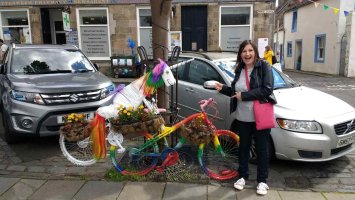 Linsey Wilson stands next to a rainbow-coloured bicycle with a paper unicorn on it. She points and smiles.