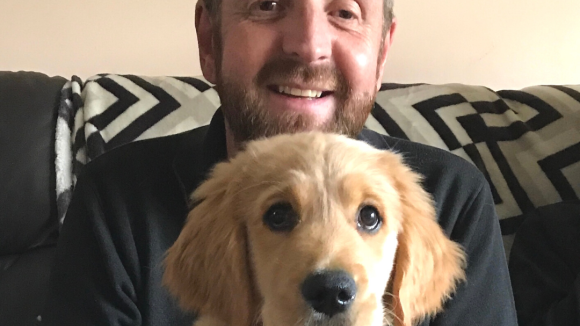 Craig sits smiling on a comfy chair with a golden retriever puppy on his lap