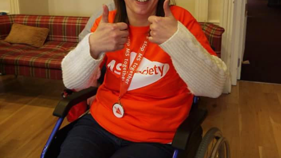 Susan Shand wearing a MS Society shirt, sat in wheelchair, smiles with thumbs-up
