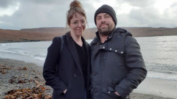 Rose and her husband standing on a beach infront of the sea