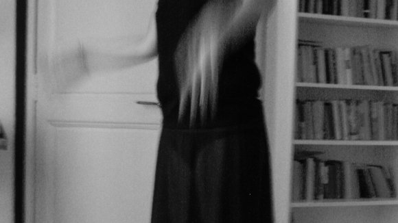 Black and white photo of Sarah doing a standing ballet pose wearing a black dress in front of a door and bookcase 