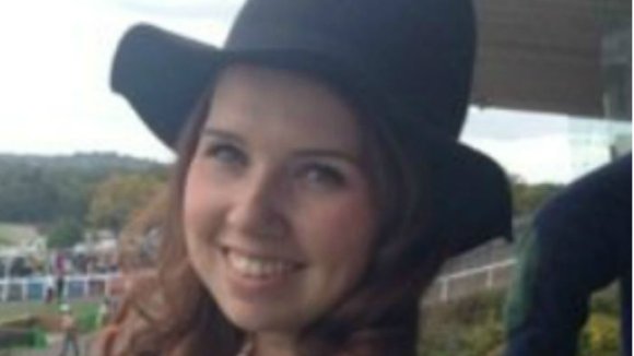 Photo shows a young woman with long brown hair and a black hat smiling at the camera.