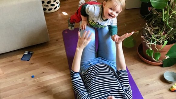 Laura, lying on a yoga mat with raised knees and hands balances baby like superman flying