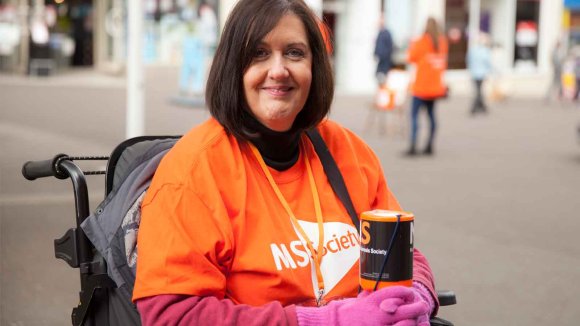 Photo shows a woman in a wheelchair in an MS Society t-shirt holding a collection tin
