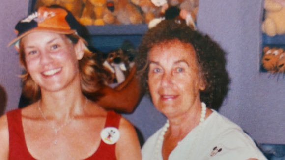 Emma as a younger woman stands smiling next to her mum, also smiling.