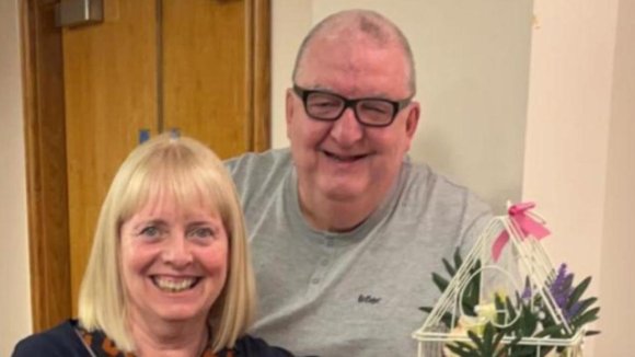 A man in a grey jumper and glasses is smiling and standing just behind a seated woman with blonde hair