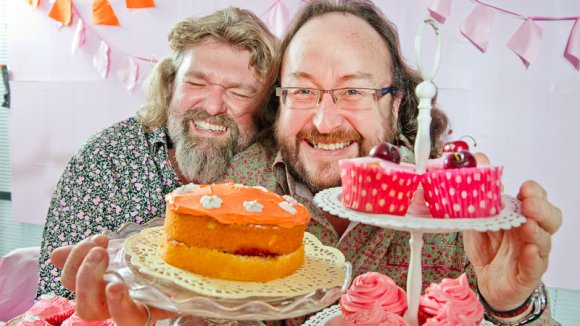 Hairy Bikers Dave and Si grinning behind a big table of cakes