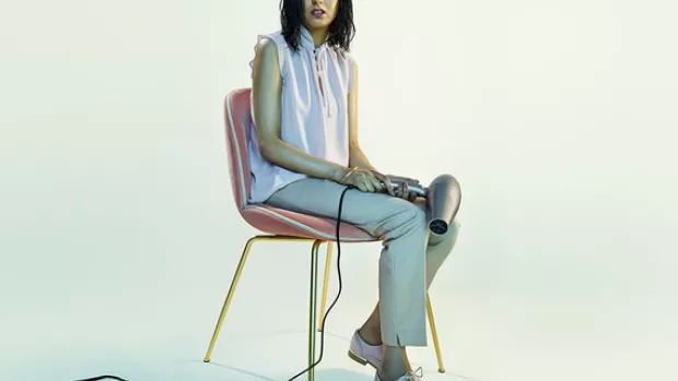 Woman with wet hair sits on a chair holding a hair dryer and looking to camera