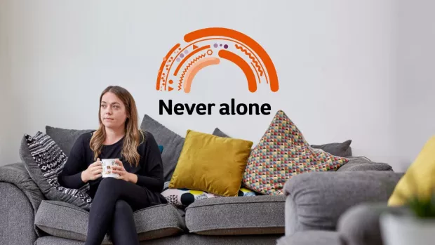 Woman sitting on a grey sofa holding a cup of tea with an orange rainbow and the words never alone behind her