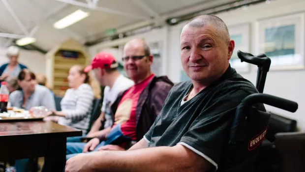 Photo shows a man in a wheelchair smiling at the camera, with his friends behind him. 
