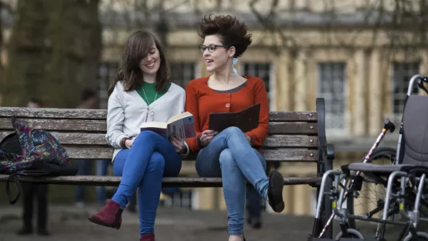 Photo shows two young women sitting on a bench, reading. 